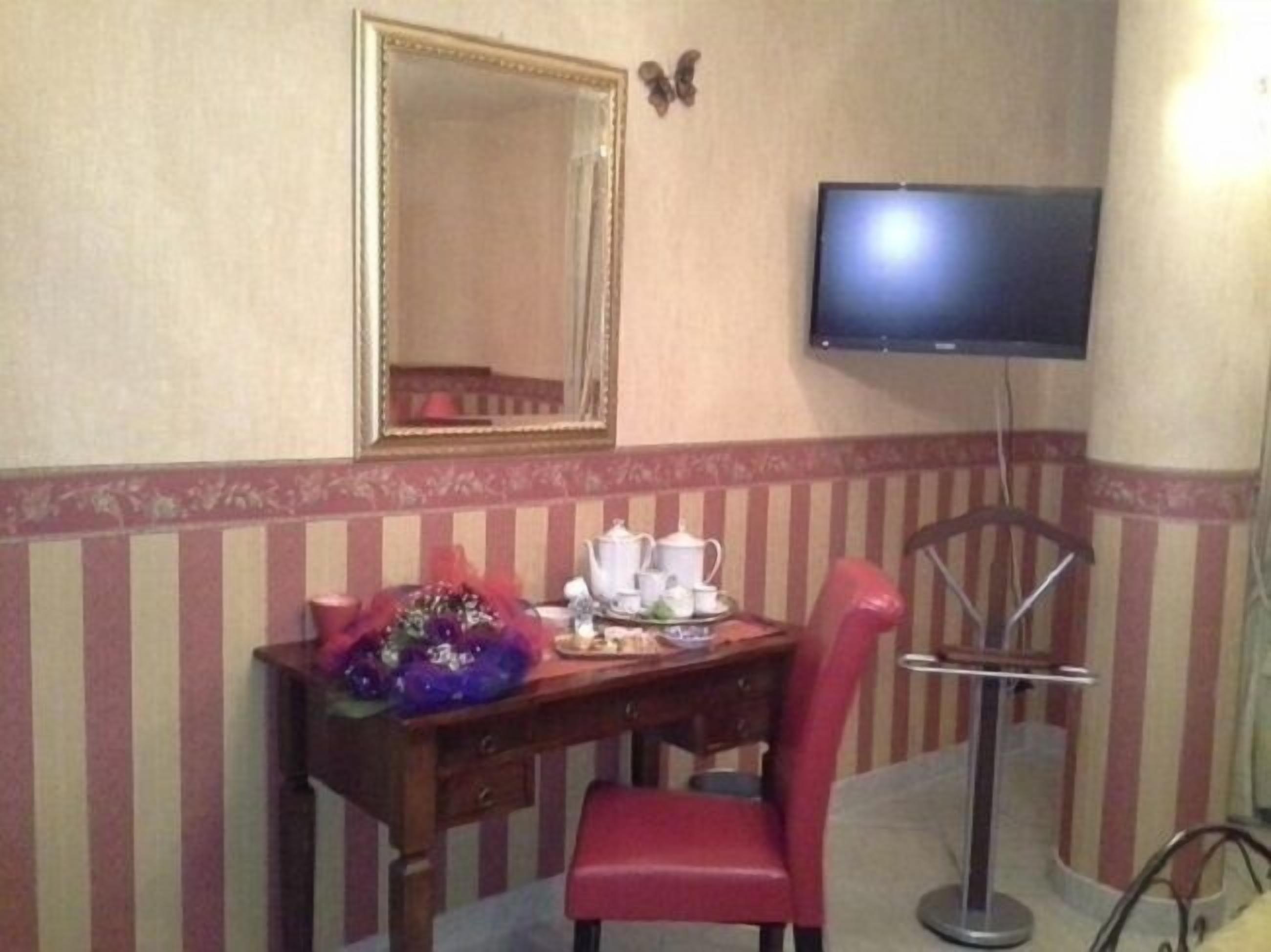 A hotel room with a wooden desk, a red chair, a flat-screen TV mounted on the wall, and a decorative mirror above the desk.