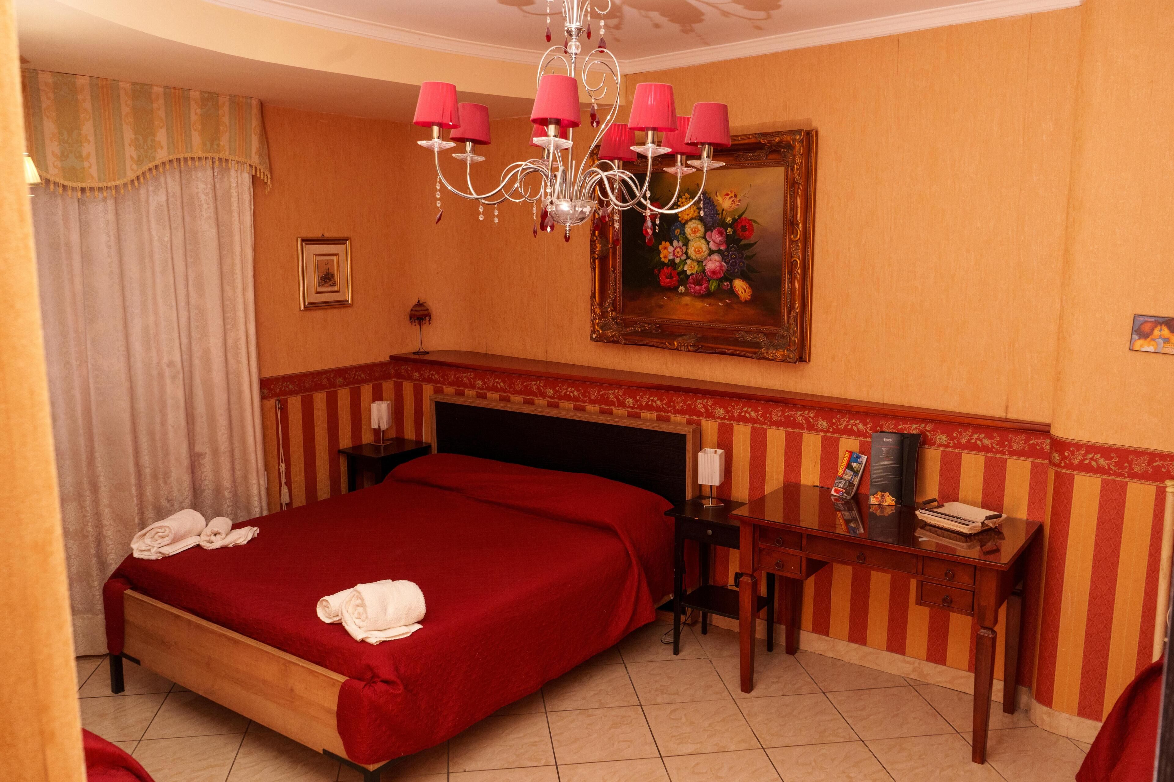 A hotel room with a large bed, a desk, a chair, a chandelier, and a painting on the wall.