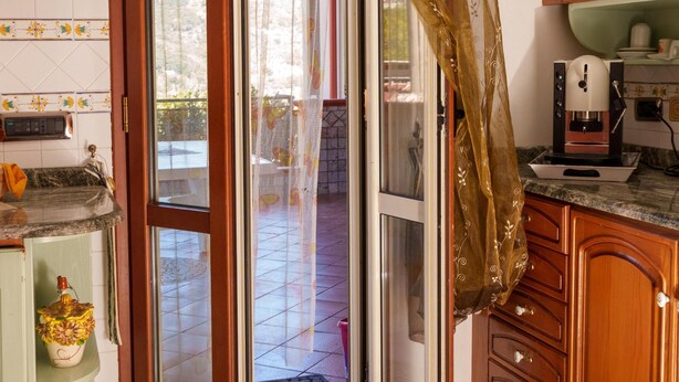 A kitchen with a sliding glass door, wooden cabinets, a coffee maker, and a green mat at the entrance. - B&B Villa Bella (Catanzaro)