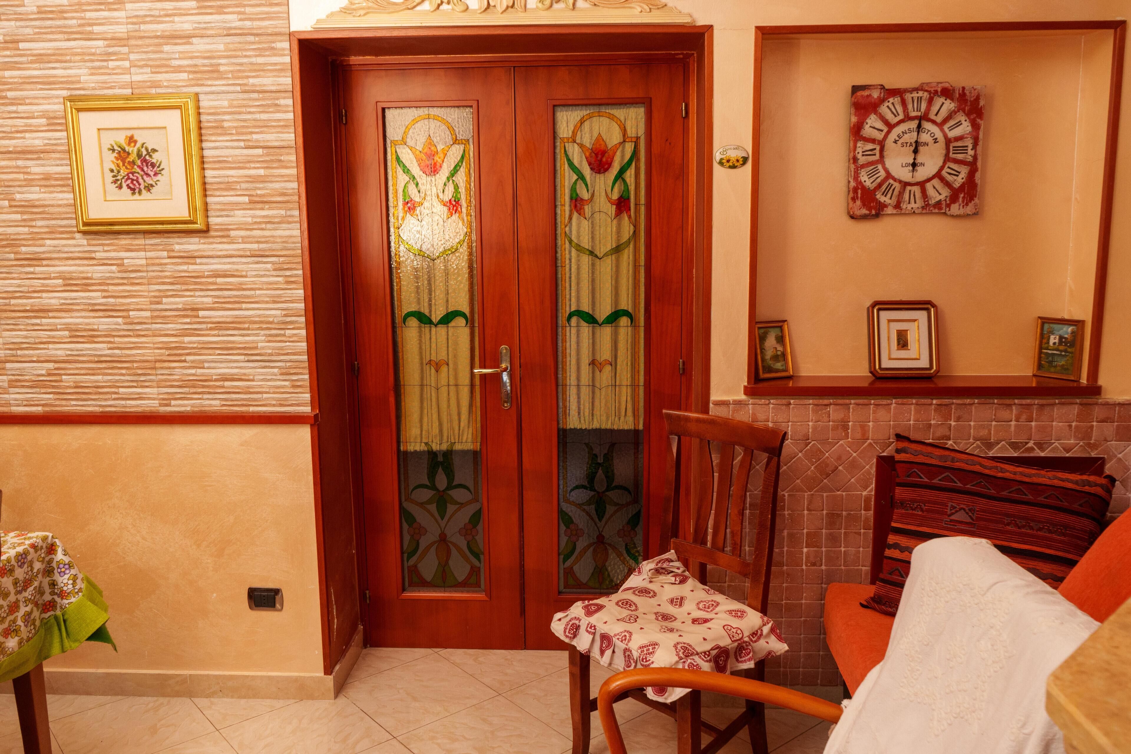 Una stanza con una porta in legno e pannelli di vetro, un orologio appeso al muro e una sedia con una tovaglia fantasia.