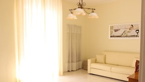 A living room with a beige sofa, a chandelier, a painting on the wall, and a window with curtains. - Leonessa di Puglia (Altamura)