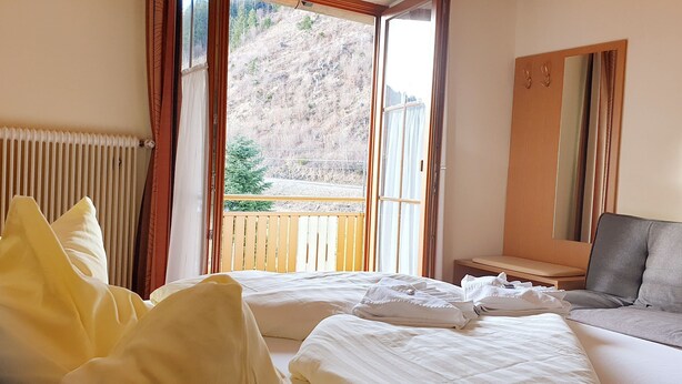 Ein Hotelzimmer mit zwei Betten, einem Schlafsofa, einem Spiegel und Blick auf einen Balkon mit Holzgeländer und einem Baum.