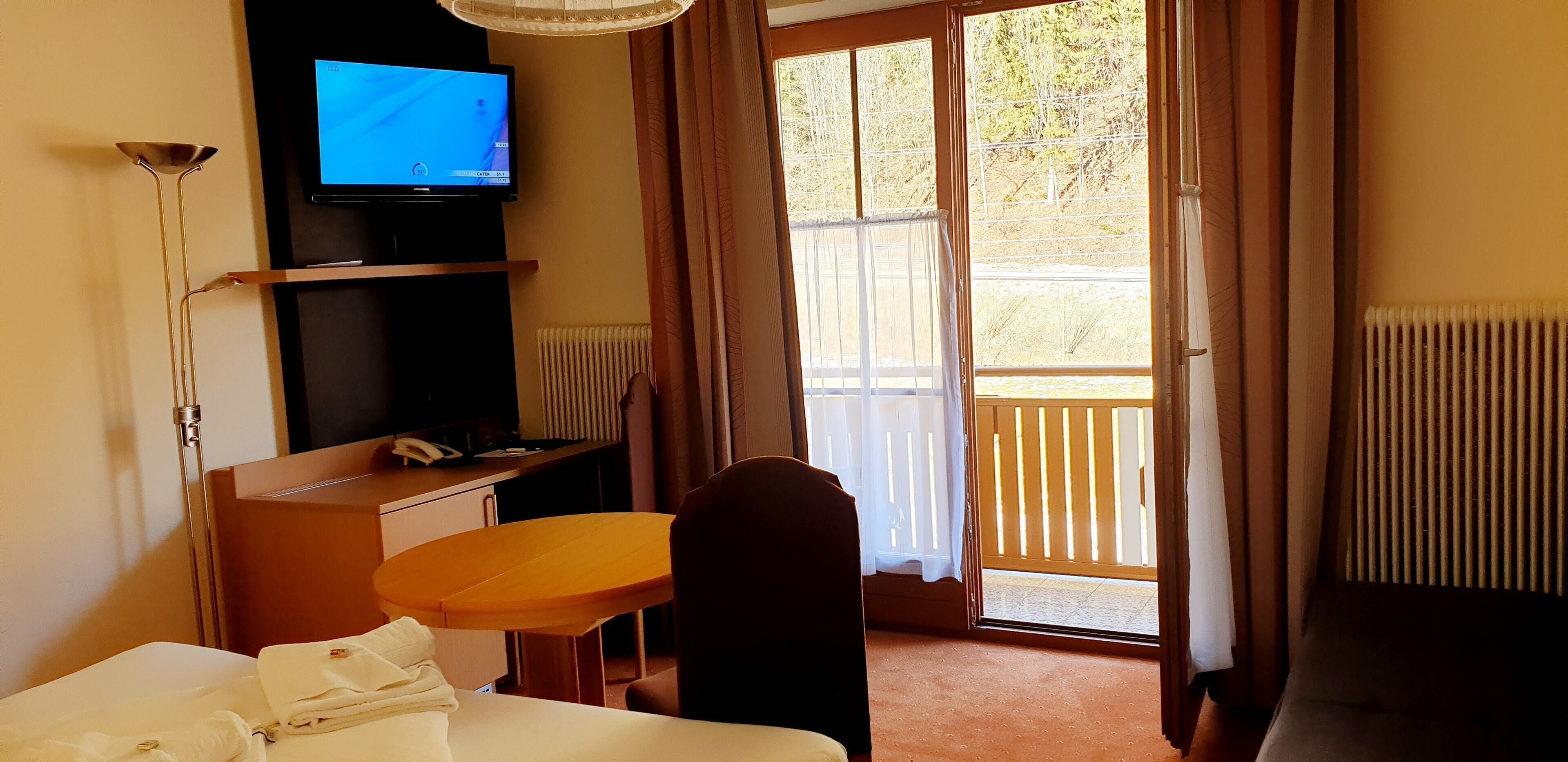 Habitación de hotel con cama, escritorio, silla, televisor y balcón con cortinas.