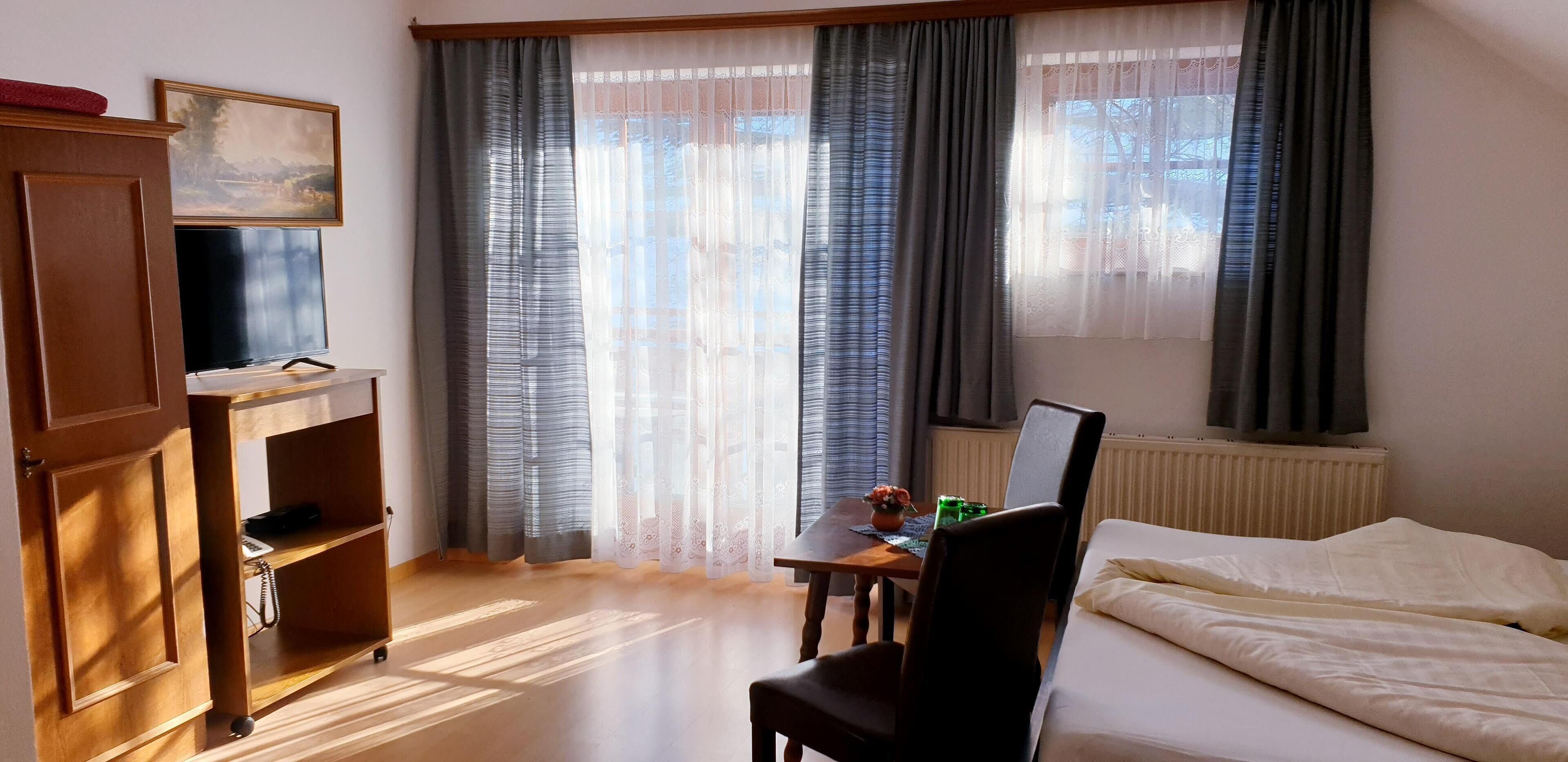 A hotel room with a bed, a desk with a computer, a TV on a stand, a wardrobe, and a window with curtains.