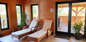Uma cabana de madeira com duas poltronas de vime com almofadas brancas, um vaso de planta e um deck de madeira visível pela porta de vidro.