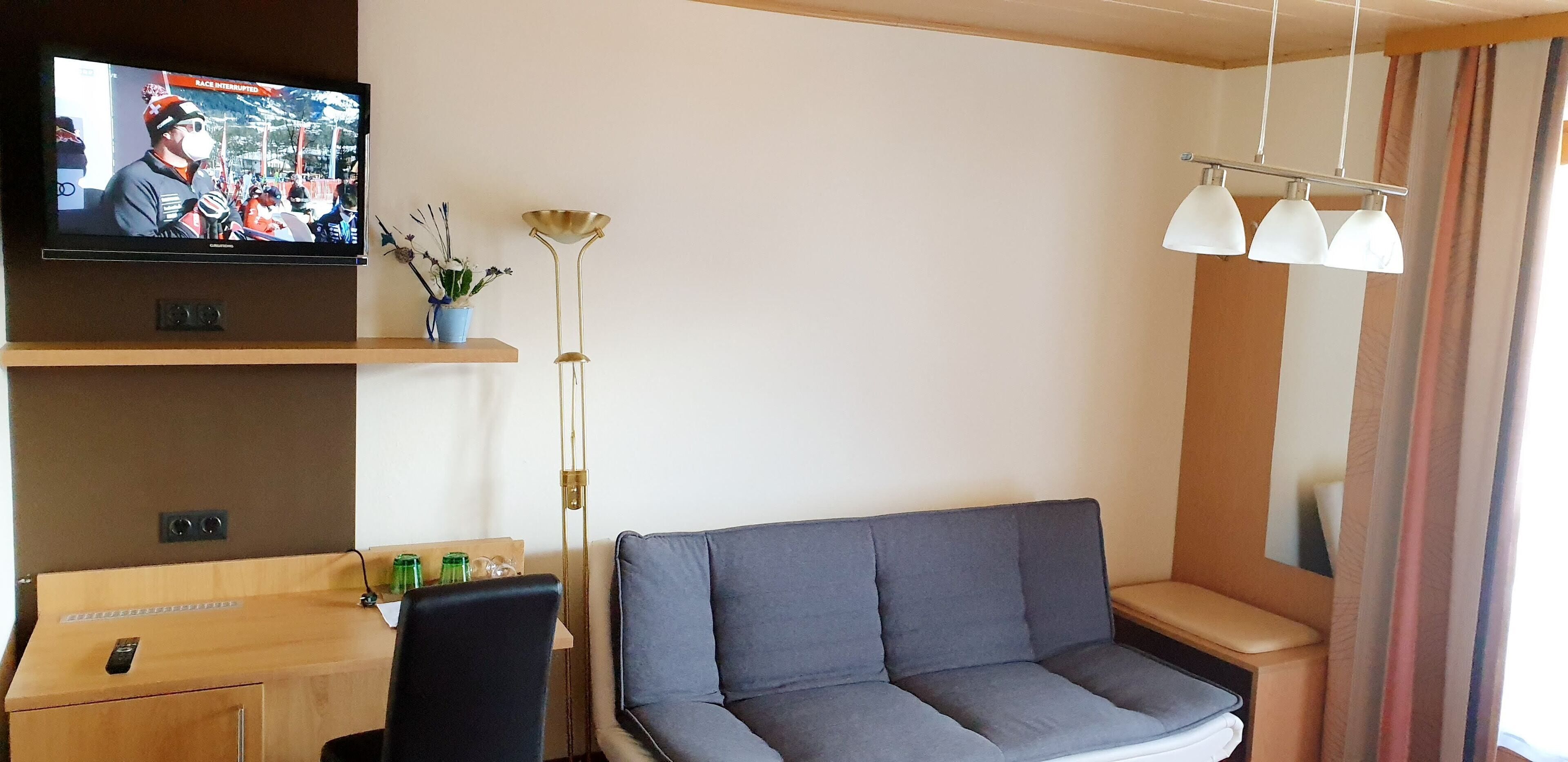 A modern hotel room with a flat-screen TV, a sofa, a desk with a chair, a floor lamp, and a window with curtains.