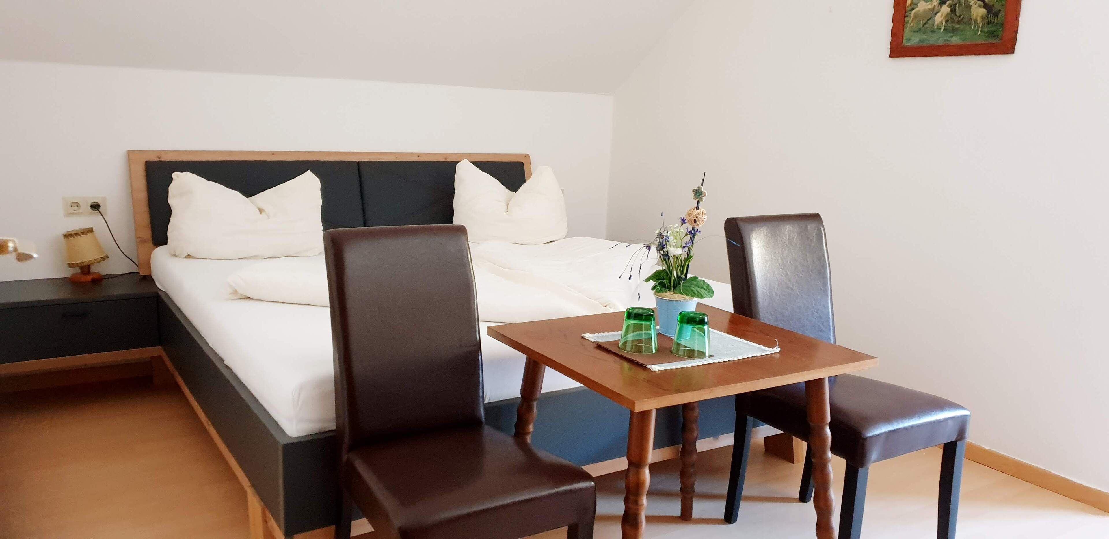 A hotel room with a bed, a wooden table with two chairs, a nightstand, and a small potted plant.