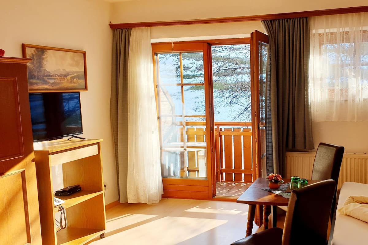 Ein Hotelzimmer mit Bett, Schreibtisch mit Stuhl, Fernseher, Fenster mit Vorhängen und Blick ins Freie.