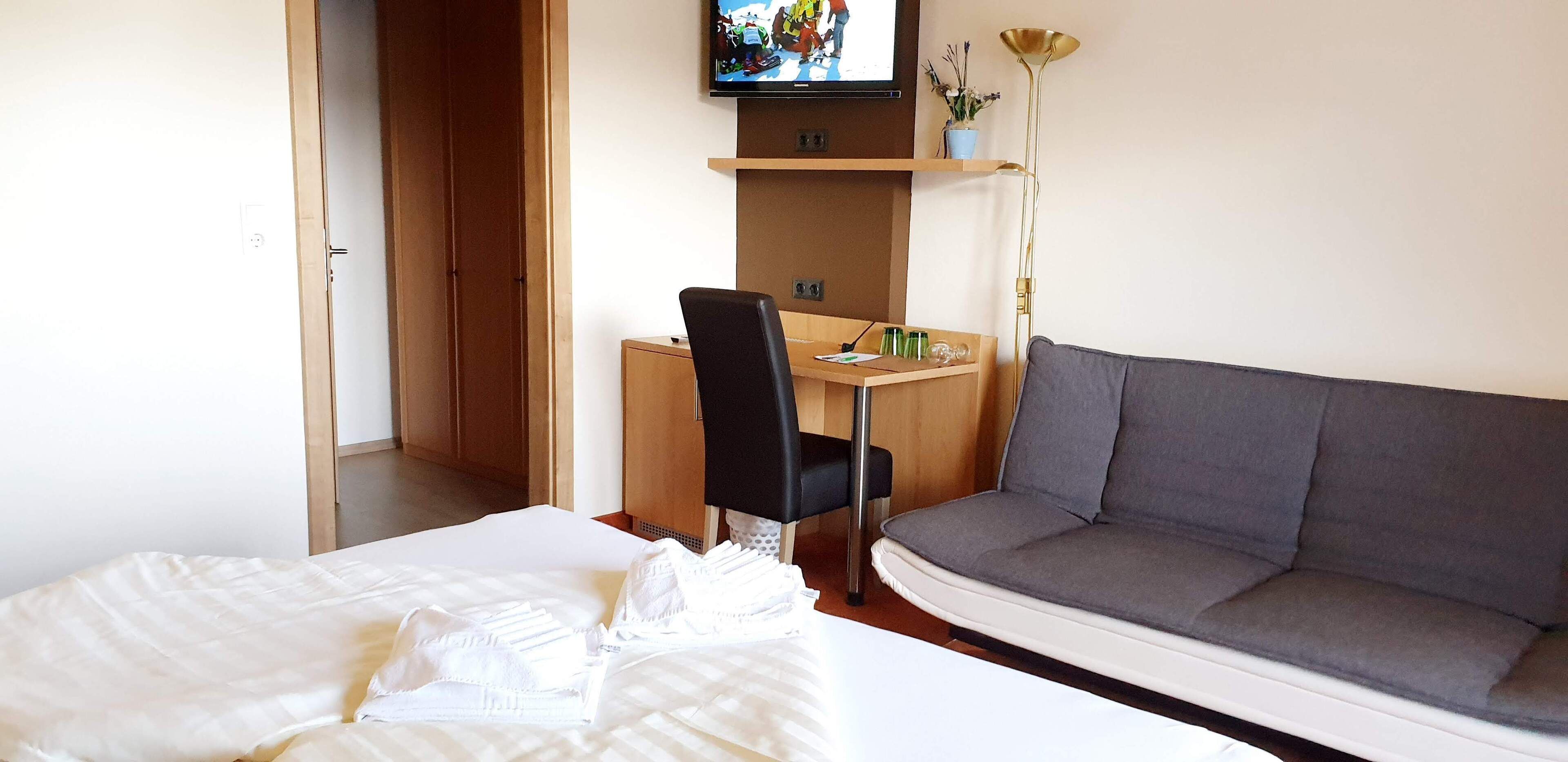 A hotel room with a bed, a gray sofa, a desk with a chair, a television mounted on the wall, and a floor lamp.