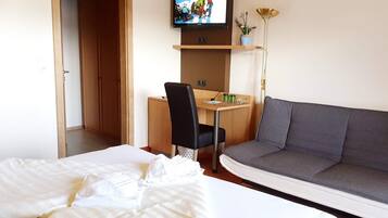 A hotel room with a bed, a gray sofa, a desk with a chair, a television mounted on the wall, and a floor lamp.