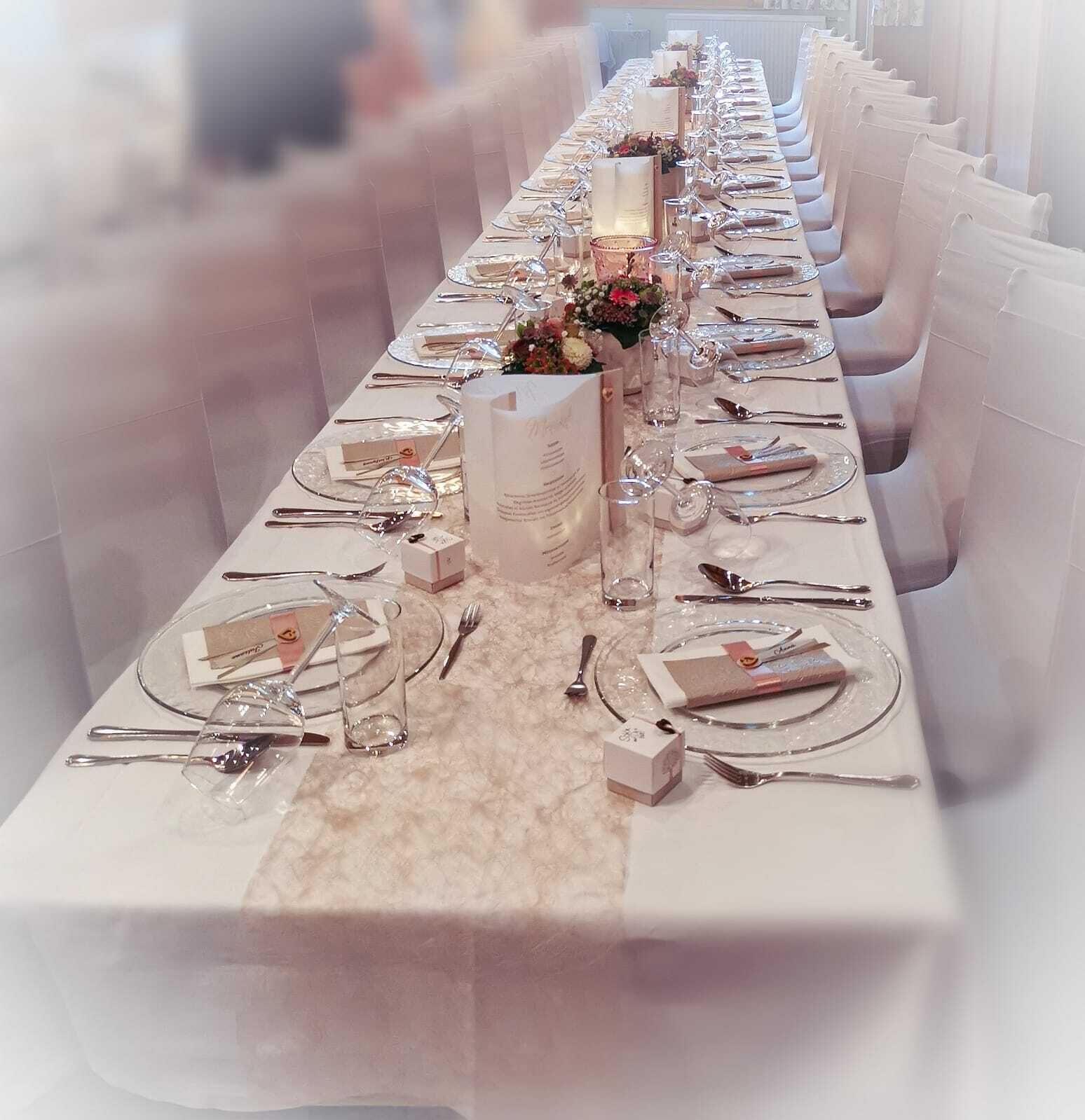 A long banquet table set with white tablecloths, silverware, and glassware, flanked by white cushioned chairs.