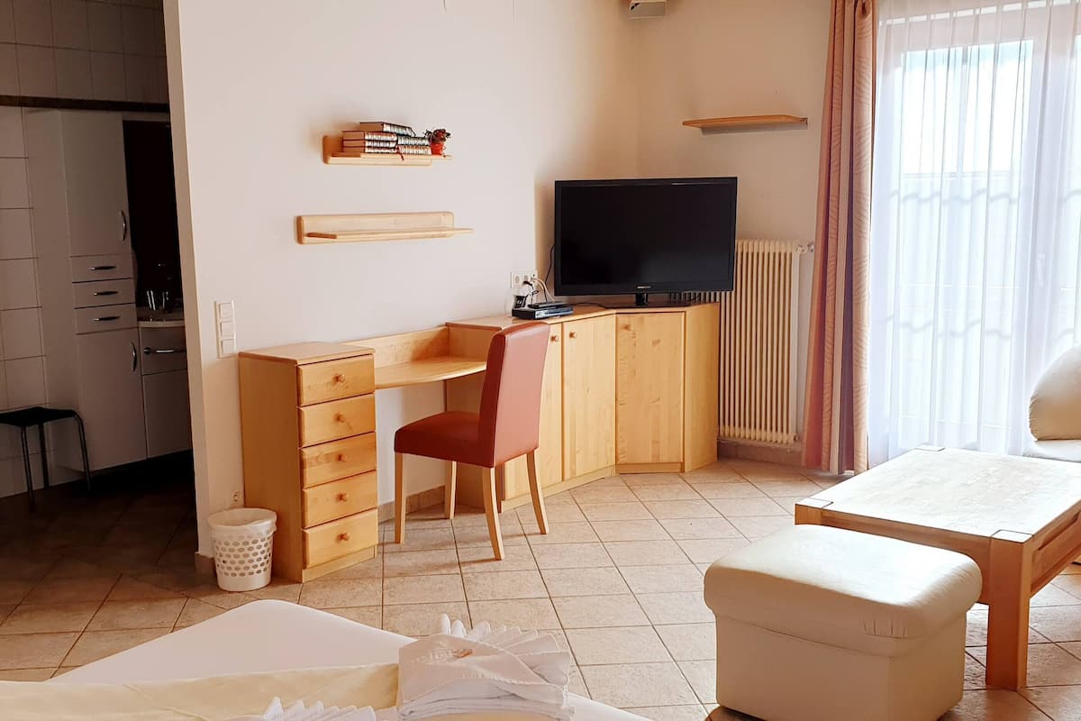 Ein kleiner, ordentlicher Raum mit einem Holztisch, einem Fernseher, einem Sofa und einem Fenster mit Vorhängen.