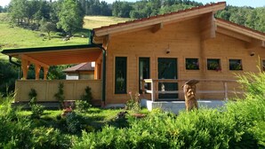Eine Holzhütte mit überdachter Veranda, einem kleinen Garten und Blick auf einen grünen Hügel.