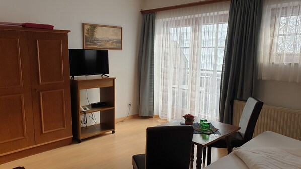 Habitación de hotel con un armario de madera, un televisor sobre un mueble bajo, una mesa con una silla y una ventana con cortinas de encaje.