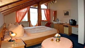 A cozy bedroom with a bed, bedside lamp, a small table with a vase, a desk with a chair, a television, and a window with curtains. - Hotel garni Schmideler (Sonthofen)