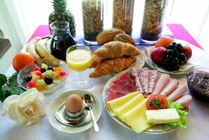 Una mesa de desayuno con cruasanes, jamón, queso, huevos y varias frutas.