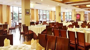 Una sala de banquets amb diverses taules rodones preparades per a dinar, grans finestres i un candelabrum.