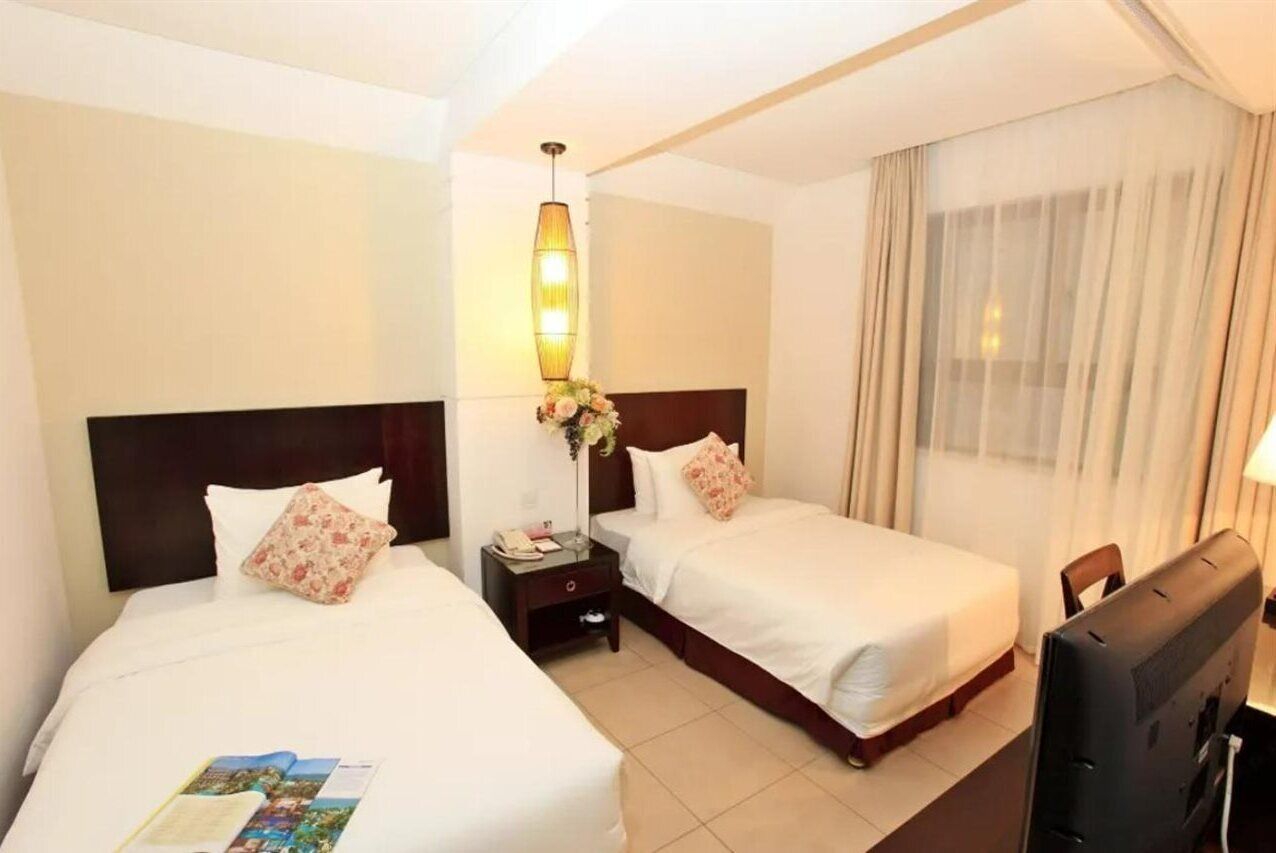 A hotel room with two beds, a TV, a bedside table with a lamp, and a window with curtains.