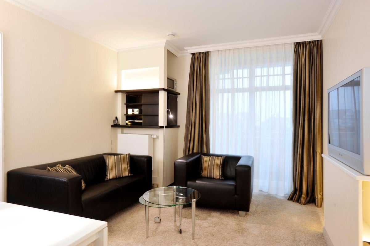 A modern living room with two black sofas, a glass coffee table, and a television on the wall.