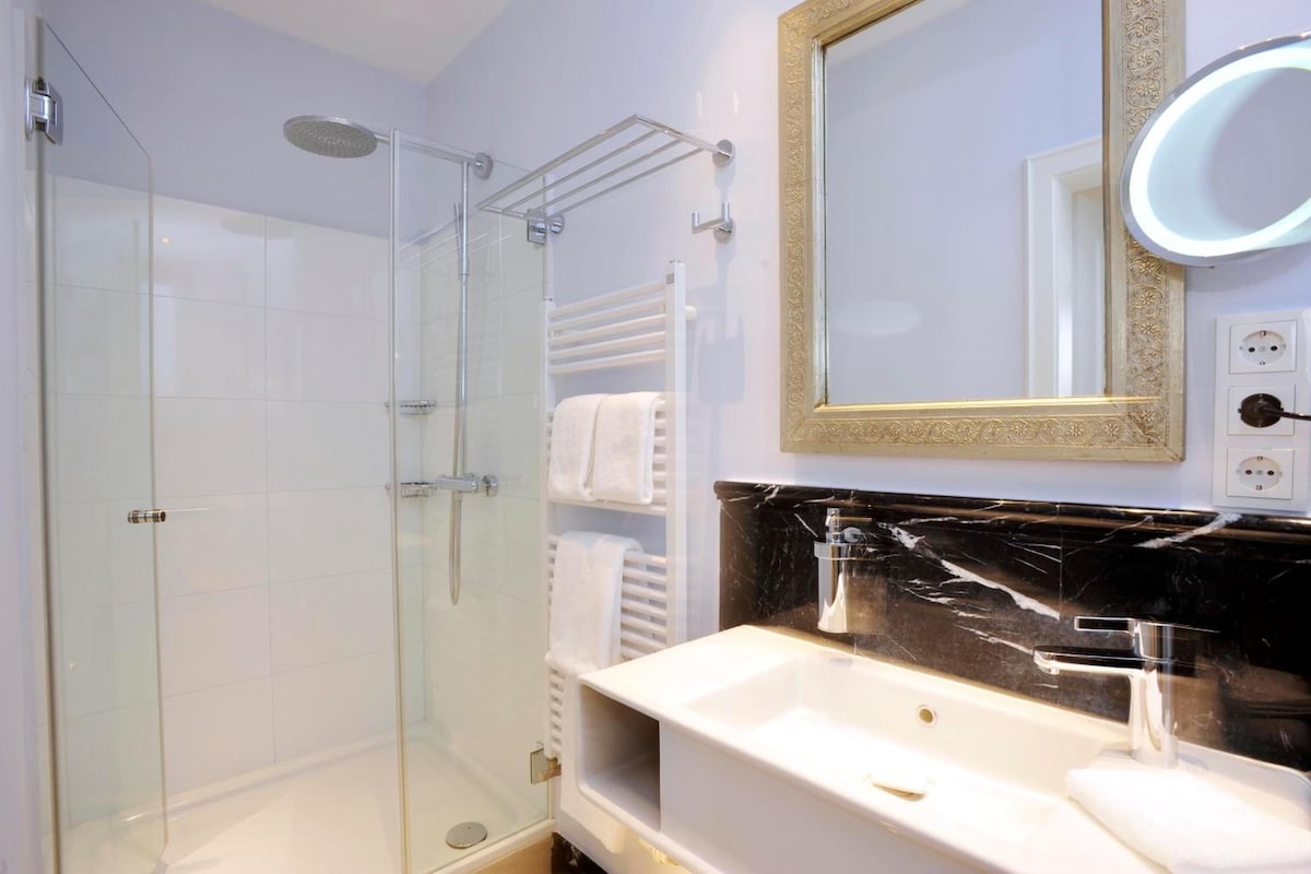 A modern bathroom with a glass shower enclosure, a vanity with a black marble countertop, and a wall-mounted mirror with a gold frame.