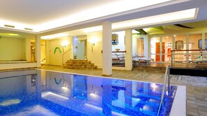 An indoor swimming pool with clear water, a sunlit area with a staircase, and a lounge with chairs.