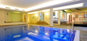 An indoor swimming pool with clear water, a sunlit area with a staircase, and a lounge with chairs.