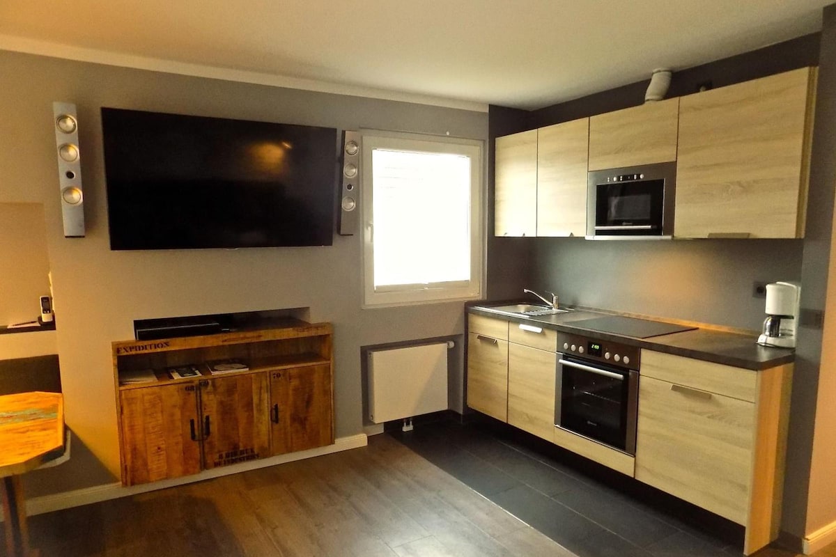 A modern kitchen with a flat-screen TV, wooden cabinets, a built-in oven, and a window providing natural light.