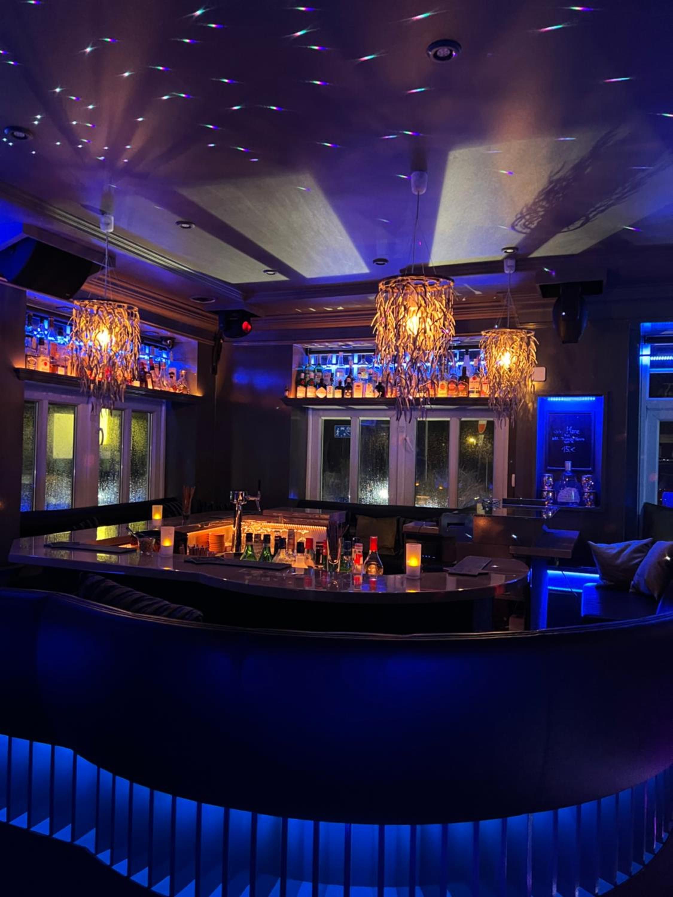 A bar area with a curved counter, multiple hanging chandeliers, and a view of the outside through large windows.