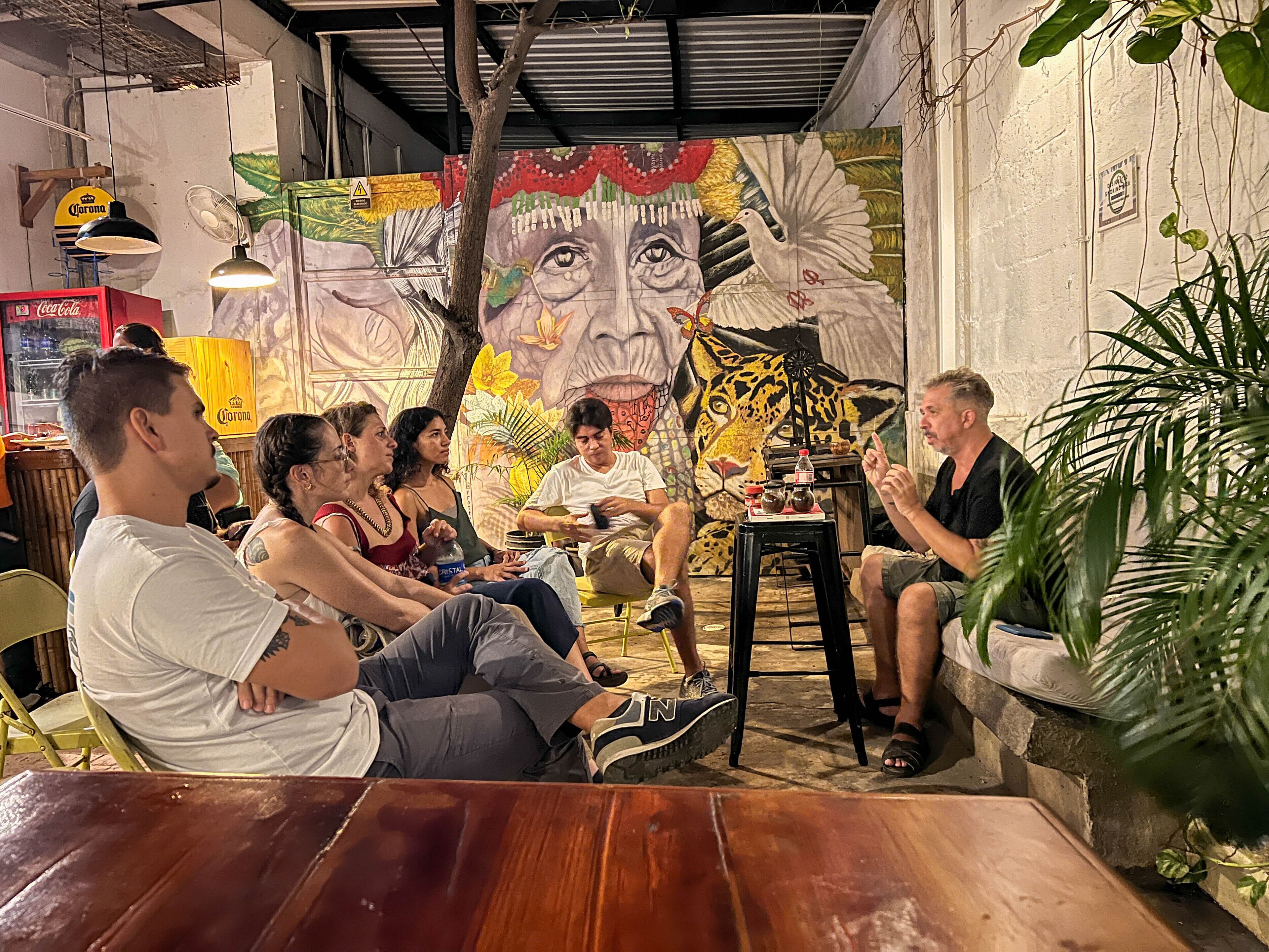 Un grupo de personas sentadas alrededor de una mesa de madera en una habitación con un mural en la pared, un refrigerador Corona y un ventilador.