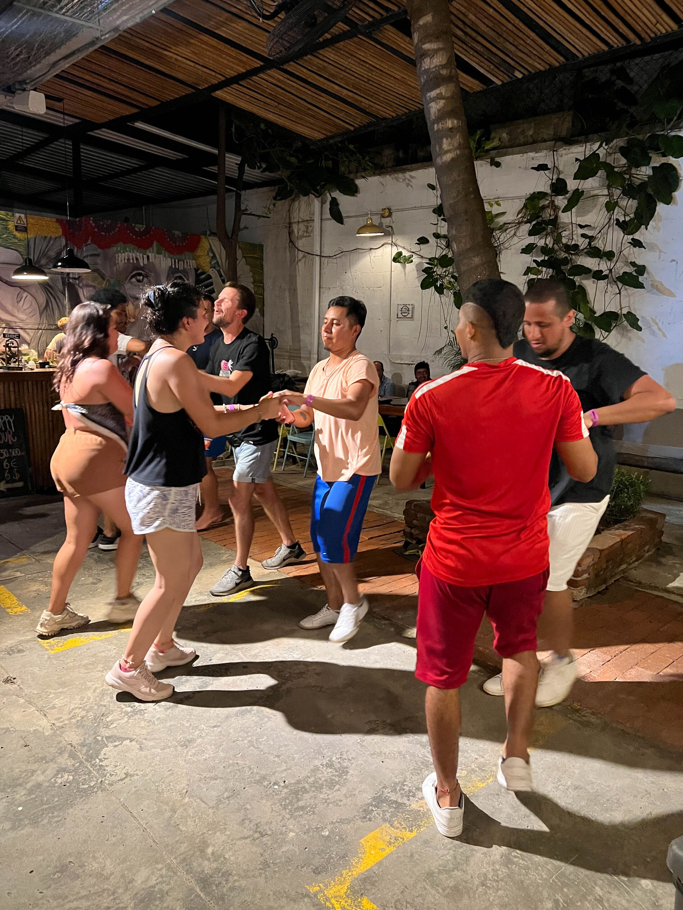 A group of people dancing in an indoor space with a concrete floor, a wooden ceiling, and a mural on the wall.
