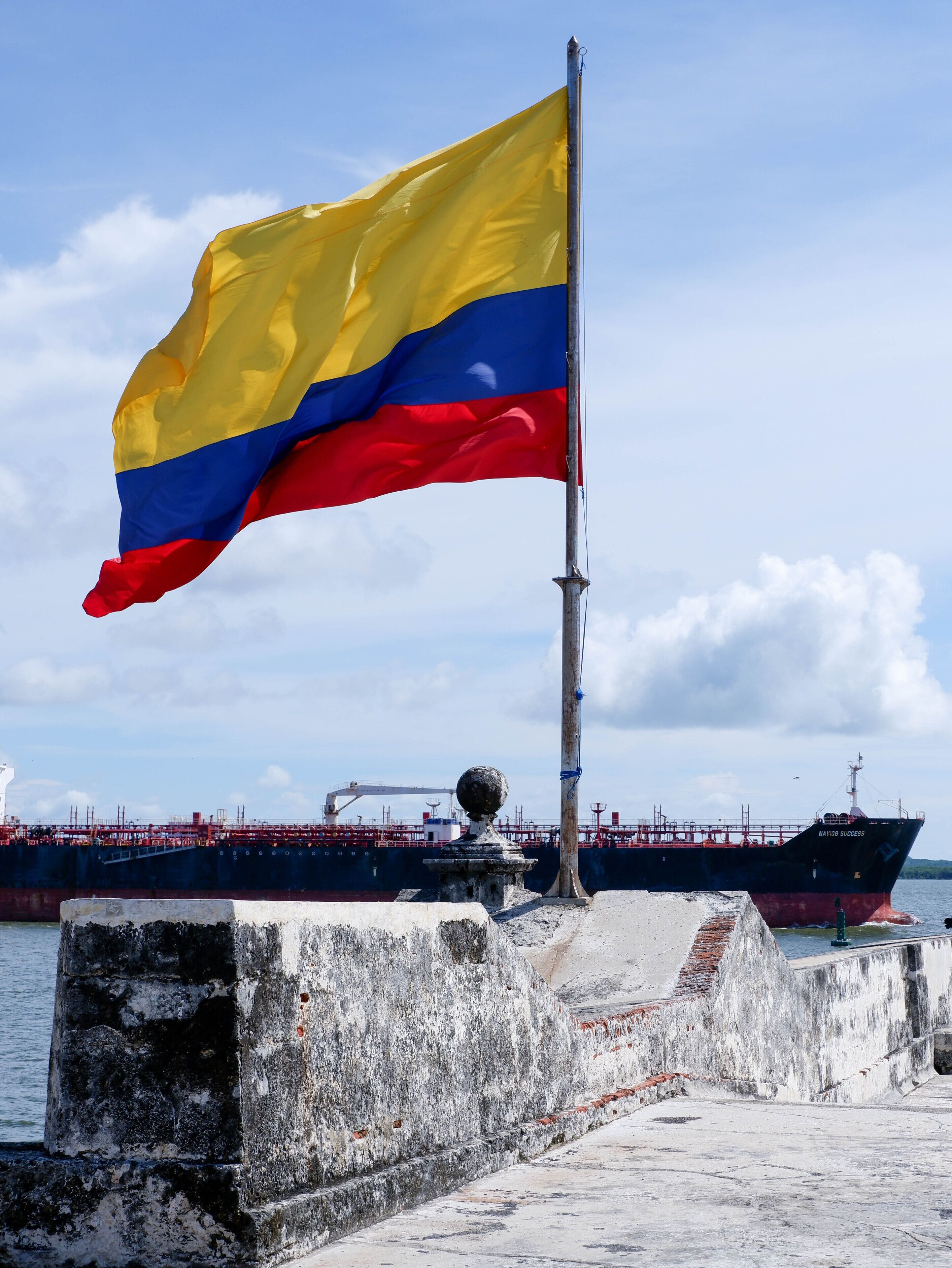 Um mastro com uma bandeira, um navio e uma estrutura de concreto à beira-mar.