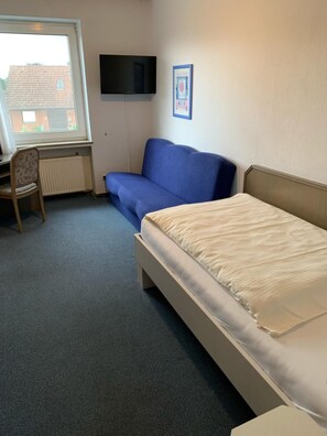 A room with a bed, a blue sofa, a chair, a radiator, a TV mounted on the wall, and a window.