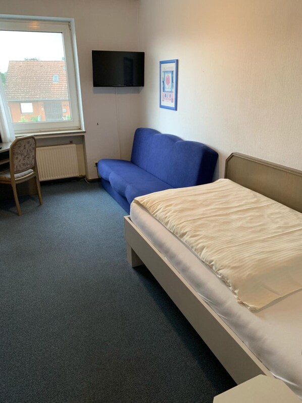 A room with a bed, a blue sofa, a chair, a radiator, a TV mounted on the wall, and a window.