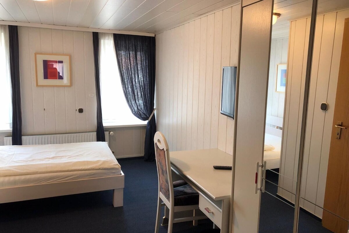 A bedroom with a bed, a desk, a chair, a wardrobe, a radiator, and a window with curtains.
