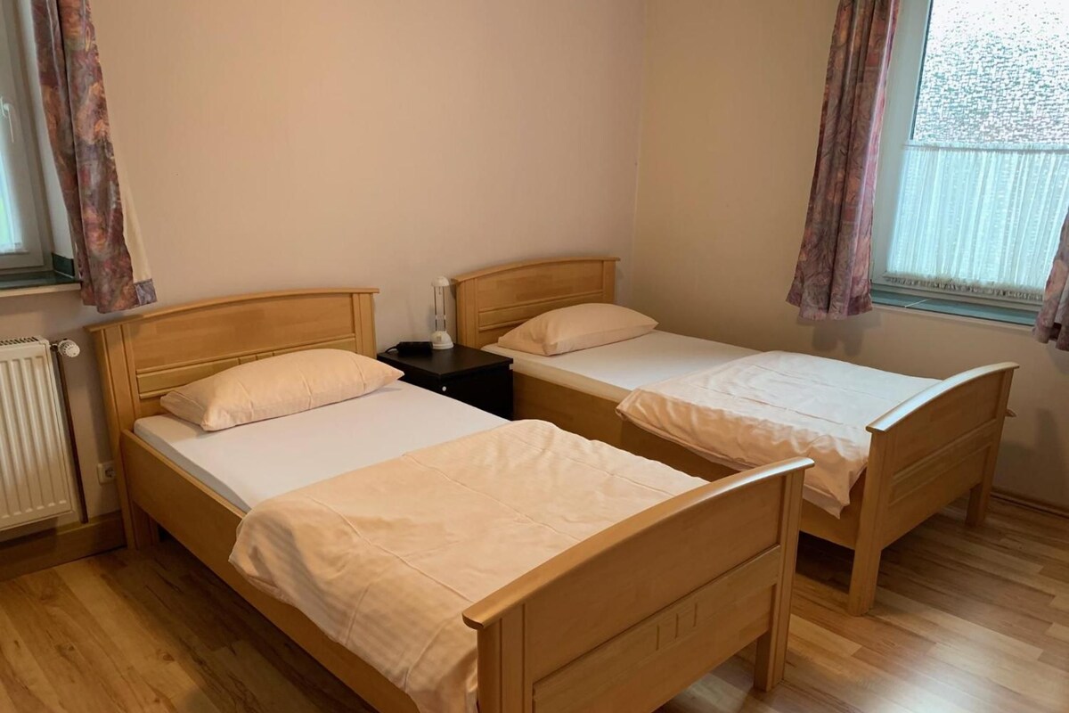 A room with two wooden beds, each with a white mattress and matching pillows. There is a nightstand with a lamp between the beds.