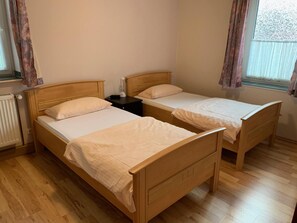 A room with two wooden beds, each with a white mattress and matching pillows. There is a nightstand with a lamp between the beds.