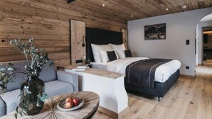 Een moderne slaapkamer met een houten plafond, een bed met wit beddengoed, een grijze bank en een ronde houten tafel met fruit.