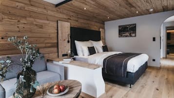 Camera moderna con soffitto in legno, un letto con biancheria bianca, un divano grigio e un tavolo rotondo in legno con frutta.