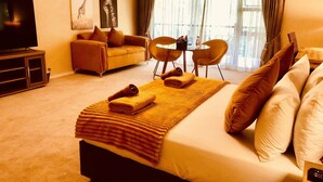 Habitación de hotel con cama, televisor, sofá, mesa de comedor con sillas y obras de arte en las paredes.