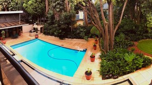 Una piscina rodeada de plantas en macetas y árboles, con una zona de estar cubierta y vista a un edificio residencial al fondo.