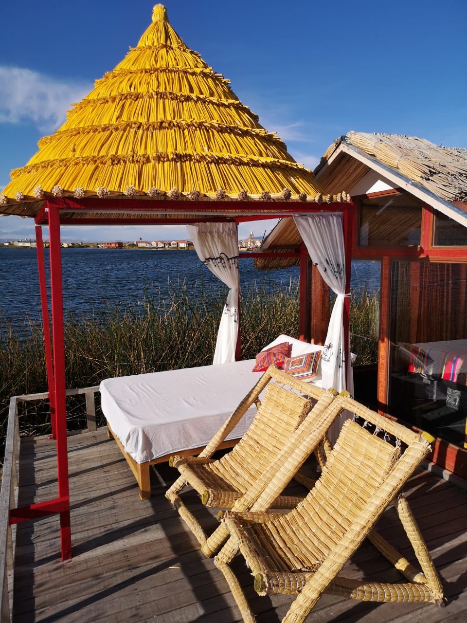 Eine Holzterrasse mit einem Bett und zwei Korbsessel, eine Struktur mit einem Reetdach und Blick aufs Wasser.