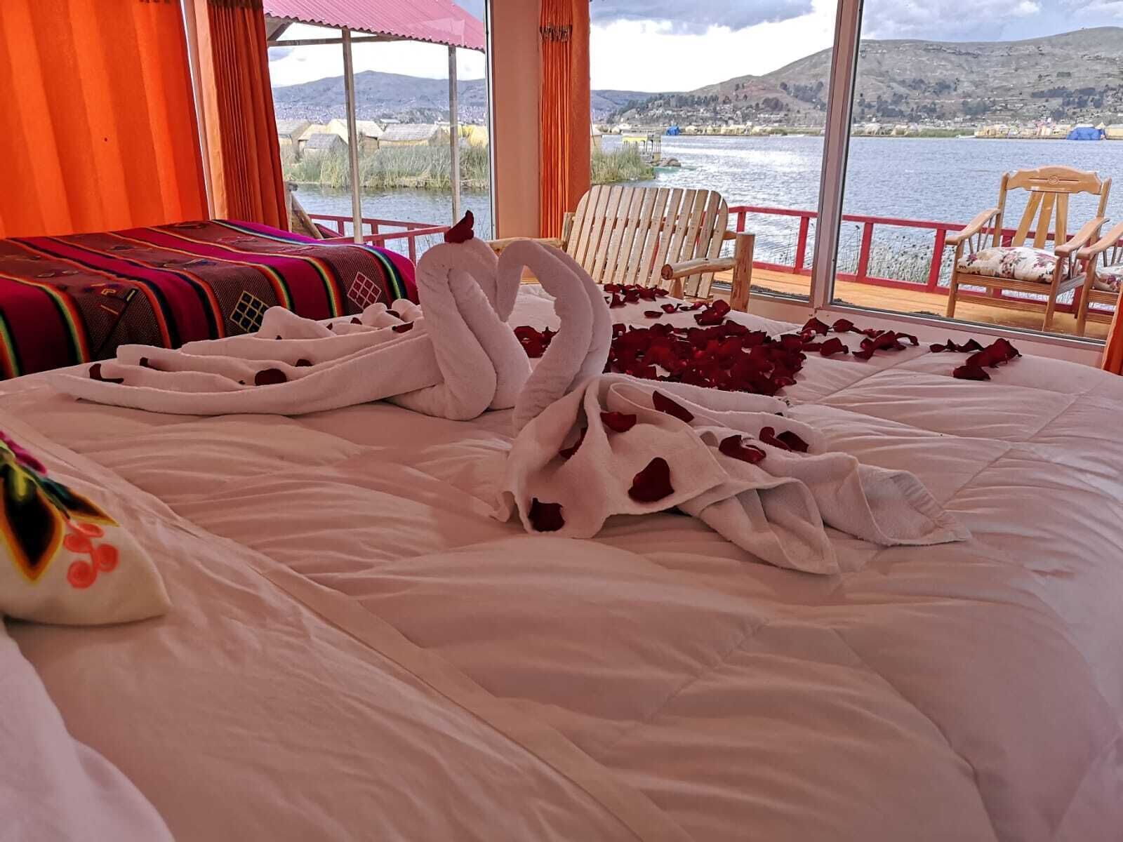 Una cama con una colcha blanca, dos cisnes hechos con toallas y pétalos de rosa esparcidos.