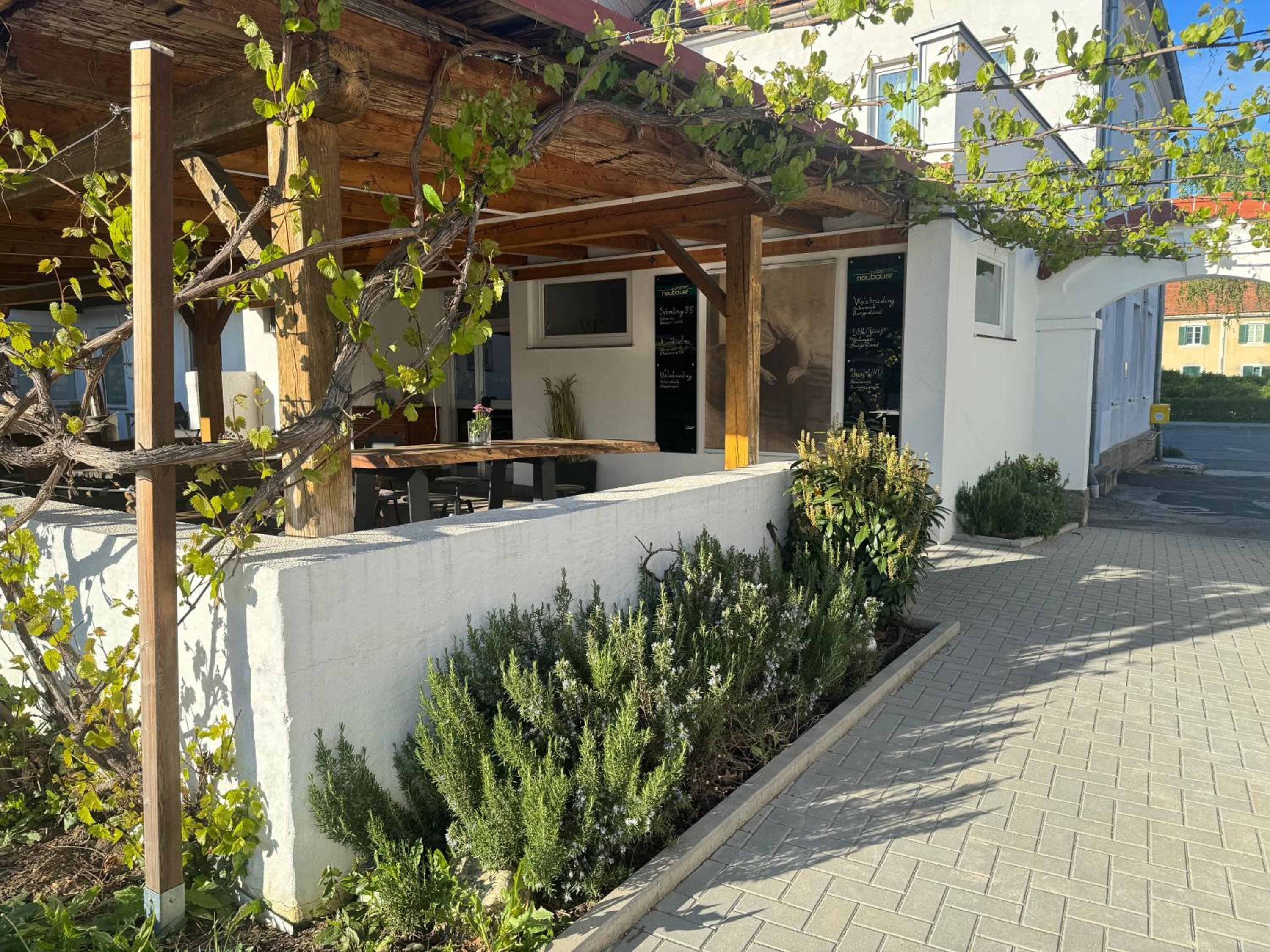 A covered outdoor seating area with a table and chairs, a white wall, and a garden with shrubs.