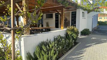 A covered outdoor seating area with a table and chairs, a white wall, and a garden with shrubs.