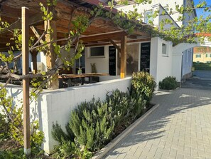 A covered outdoor seating area with a table and chairs, a white wall, and a garden with shrubs.