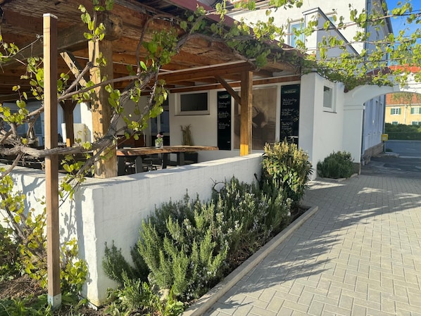 Un espace extérieur abrité, avec une table et des chaises, un mur blanc et un jardin agrémenté d’arbustes.