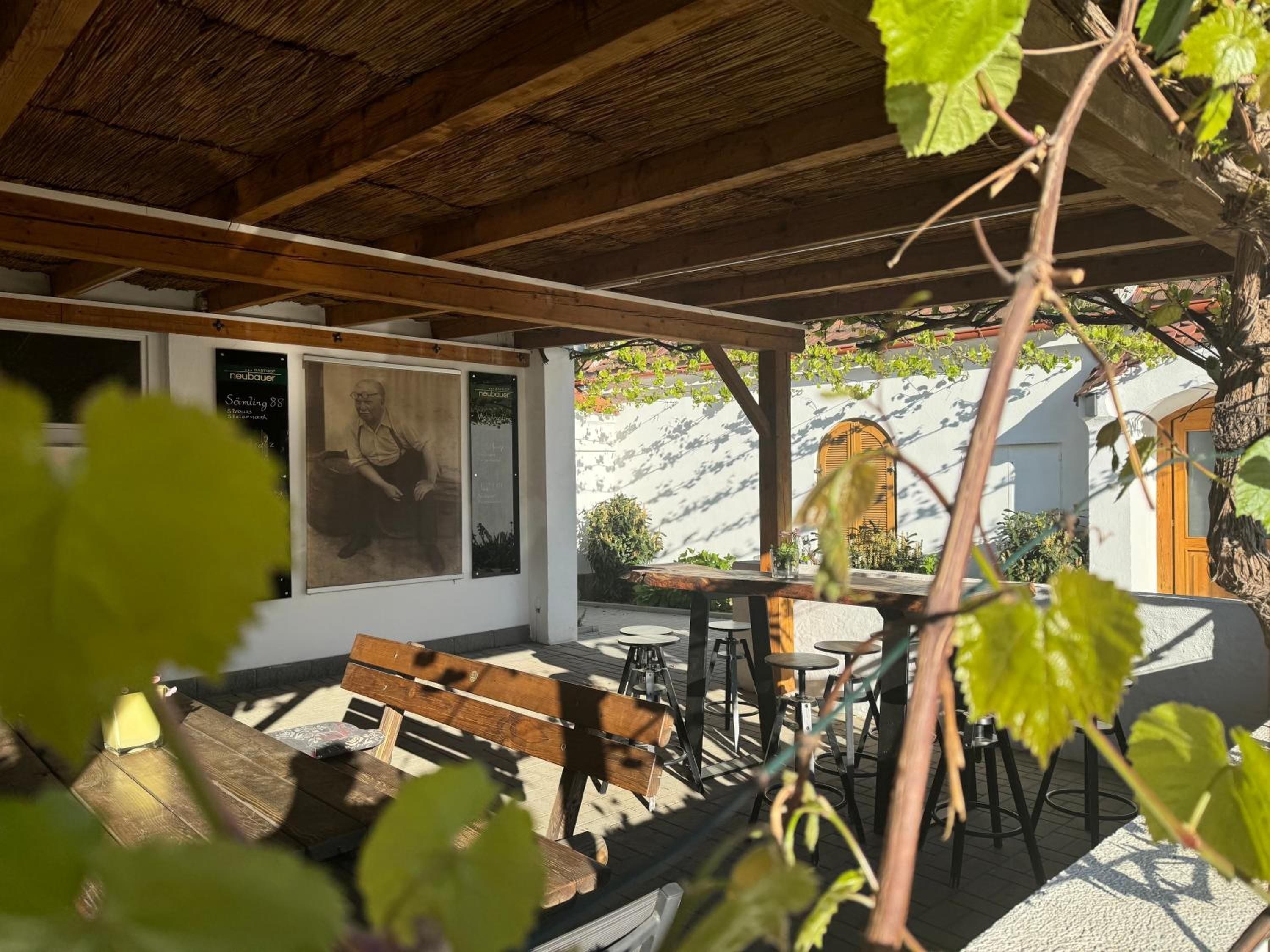 An outdoor seating area with wooden benches and tables, a pergola overhead, and a wall with a poster.