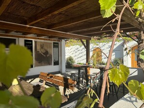 An outdoor seating area with wooden benches and tables, a pergola overhead, and a wall with a poster.