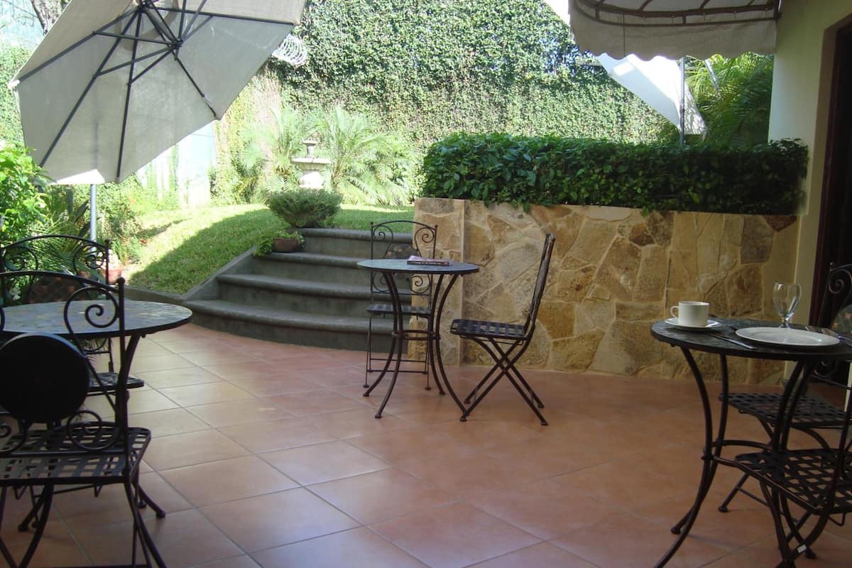 An outdoor patio area with tiled flooring, metal tables and chairs, a stone wall, and a small garden with steps leading up to a stone fountain.