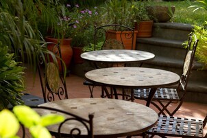 Un patio con dos mesas redondas y sillas de metal, rodeado de plantas en macetas y escalones que llevan a un jardín.
